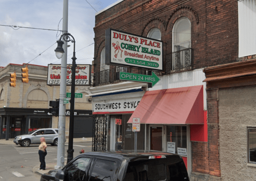 Sign for Duly's Place, a 24-hour breakfast spot, on a brick building with nearby shops and a street view.
