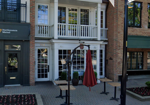 A brick building with large windows, outdoor seating, and a red umbrella, located in a quaint urban area.