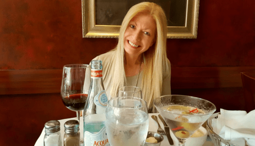 A smiling woman with long blonde hair sits at a table set with drinks, including wine and a cocktail, in a restaurant.
