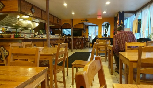 Cozy restaurant interior with wooden tables and chairs, featuring a kitchen area and a few patrons. Warm, inviting atmosphere.