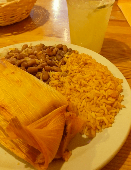 A plate with a tamale, rice, and beans, accompanied by a glass of lemonade.