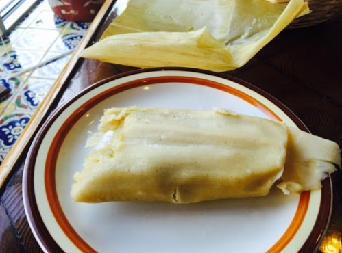 A tamale on a plate, partially wrapped in corn husk, with chopsticks and additional husks in the background.