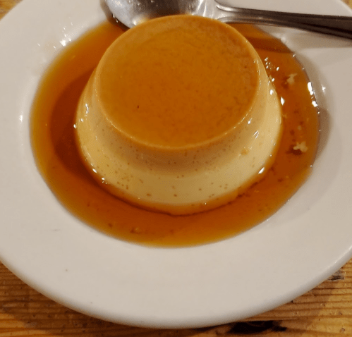 A smooth caramel flan on a white plate, surrounded by a glossy caramel sauce, with a spoon resting nearby.