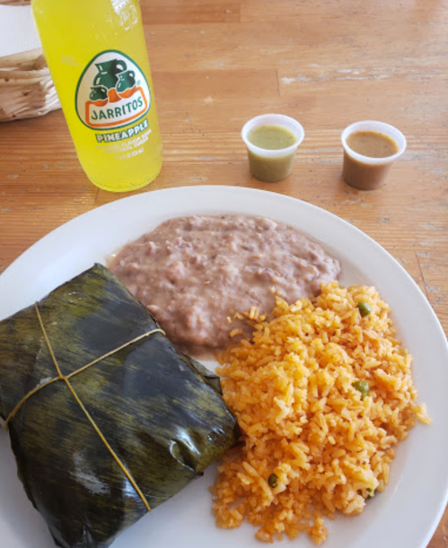 A plate with rice, refried beans, a wrapped tamale, and two sauces, alongside a bottle of pineapple Jarritos.