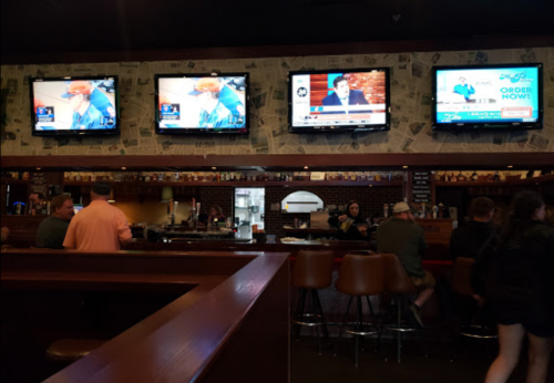 A bar interior with multiple TVs showing news and sports, patrons seated at the bar, and a cozy atmosphere.