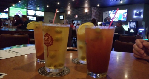 Four colorful cocktails with garnishes sit on a bar table, with a lively sports bar atmosphere in the background.