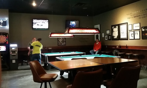 A dimly lit bar with pool tables, a dartboard, and patrons watching TV and playing games.