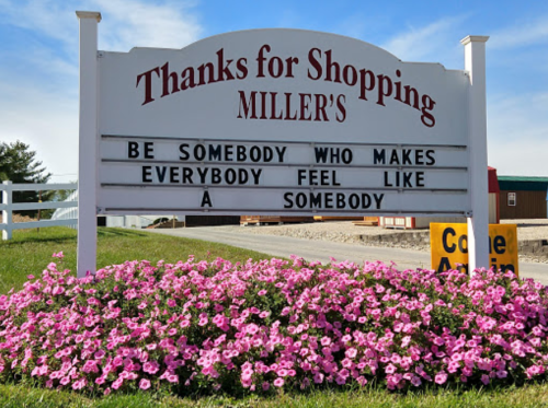 Sign at Miller's with a message encouraging kindness: "Be somebody who makes everybody feel like a somebody." Surrounded by flowers.