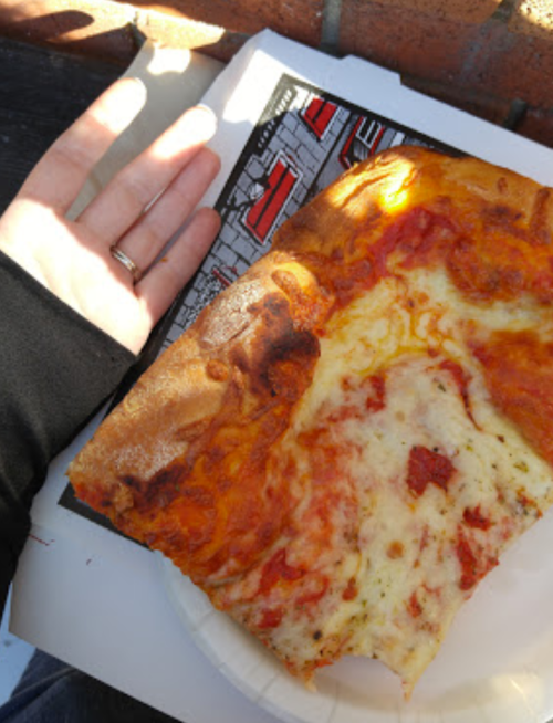 A hand next to a large slice of pizza on a paper plate, with a partially eaten corner and a colorful background.