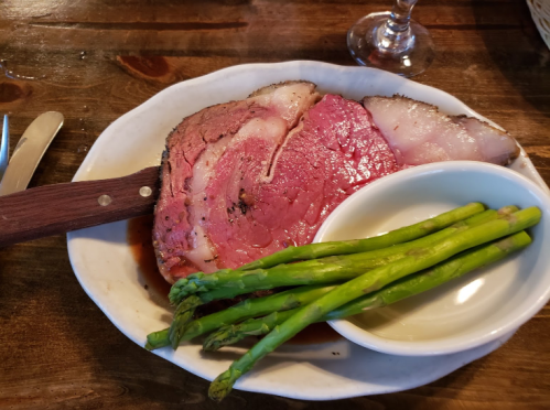 A plate of sliced prime rib with a side of asparagus and a small bowl of sauce on a wooden table.