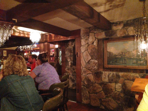 Cozy bar interior with stone walls, wooden beams, and patrons seated at the counter, illuminated by hanging lights.