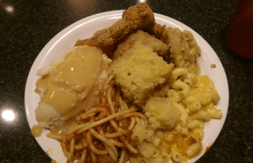 A plate of comfort food featuring mashed potatoes with gravy, fried chicken, macaroni and cheese, spaghetti, and cornbread.