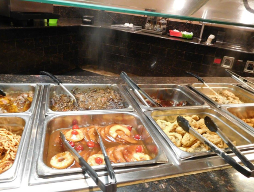 A buffet with various dishes in metal trays, including meats, vegetables, and garnishes, with steam rising.