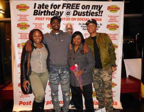 A group of four people poses together in front of a promotional banner for Dusties restaurant, celebrating a birthday.