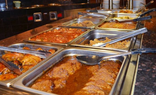 A buffet spread featuring various dishes in metal trays, including meats and sides, with serving utensils.