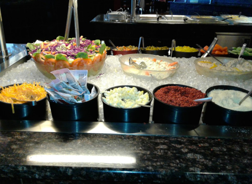 A salad bar with various toppings in black bowls, including cheese, vegetables, and a large bowl of salad.