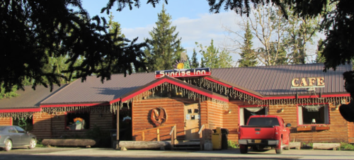 A rustic log cabin cafe with a sign reading "Sunrise Inn," surrounded by trees and a clear blue sky.
