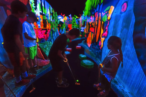 Children playing mini-golf in a vibrant, neon-lit indoor course with glowing decorations.
