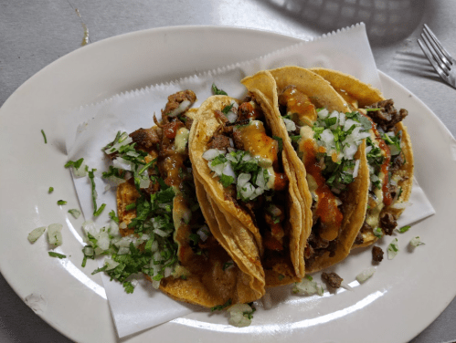 Three tacos filled with meat, topped with salsa, onions, and cilantro, served on a white plate.