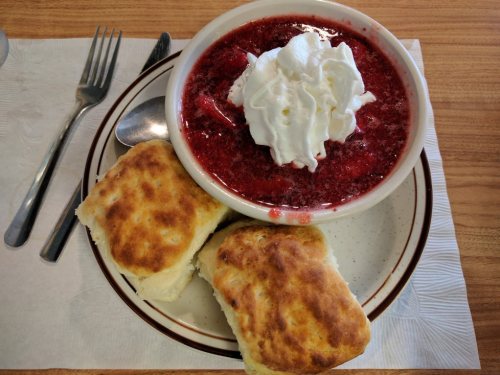 A bowl of strawberry dessert topped with whipped cream, served with two flaky biscuits on a plate.