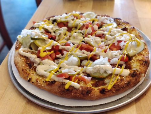 A pizza topped with pickles, tomatoes, onions, and drizzled with mustard and sauce on a sesame seed crust.