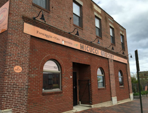 Brick building with large windows and a sign reading "Micucci Grocery," featuring Italian food offerings.