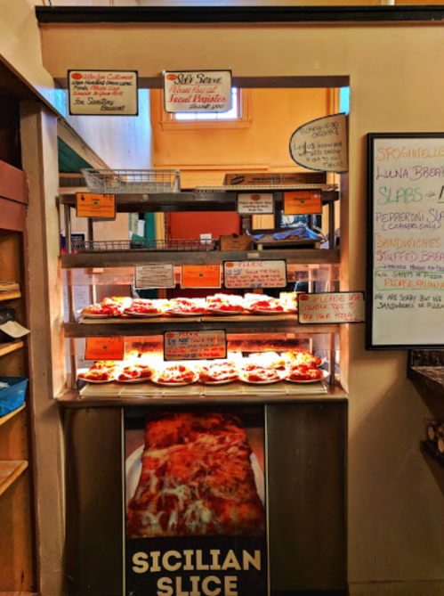 A display case filled with various pizzas, including a large Sicilian slice, in a cozy pizzeria setting.