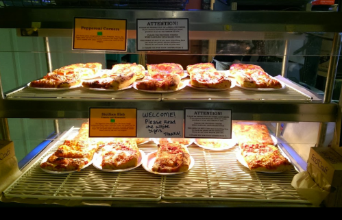 A display case with various pizza slices, including pepperoni and Sicilian, with signs for customer instructions.