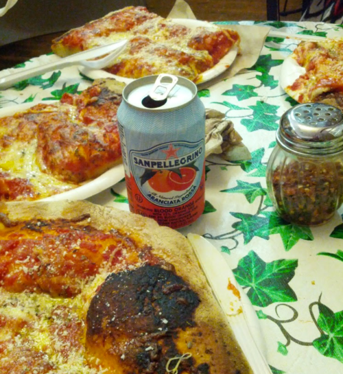 A table with pizza slices, a can of San Pellegrino, and condiments on a patterned tablecloth.
