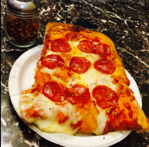 A slice of pepperoni pizza with melted cheese on a paper plate, next to a shaker of red pepper flakes.