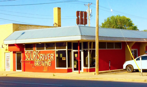 Exterior of Sour River Brewing, featuring a red facade, large windows, and a vintage-style sign. Clear blue sky above.