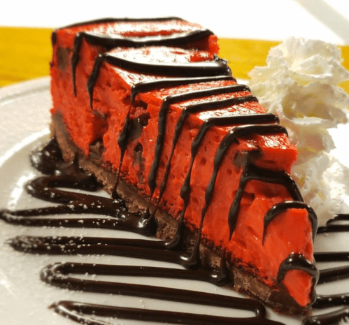 A slice of vibrant red cheesecake drizzled with chocolate sauce, served with a dollop of whipped cream on a white plate.