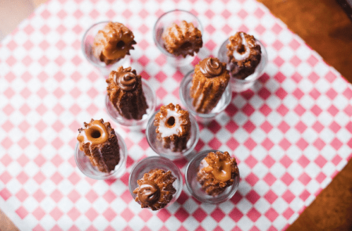 A top-down view of mini churros in shot glasses, topped with chocolate and caramel sauces on a checkered tablecloth.