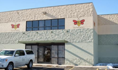 Exterior of a winery building with a bee logo, large windows, and a green awning. A parked vehicle is in front.