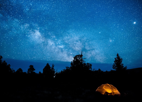 A glowing tent under a starry night sky filled with the Milky Way and silhouetted trees.