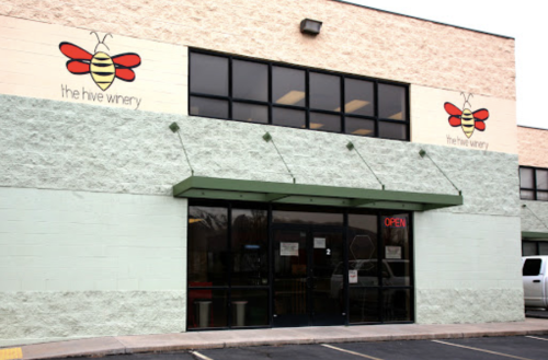 Exterior of The Hive Winery, featuring a green facade, large windows, and a sign with a bee logo. The door is open.