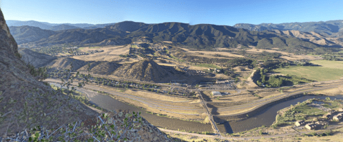 A panoramic view of a valley with a river, mountains, and fields under a clear blue sky.