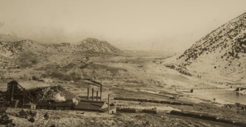 Black and white landscape of a valley with mountains, industrial buildings, and a train near a river.
