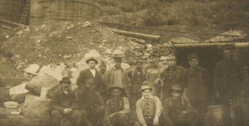 A group of men in historical clothing poses near a mining site, surrounded by rocks and equipment.