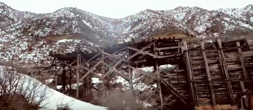 A weathered wooden bridge stands against a backdrop of snow-covered mountains.