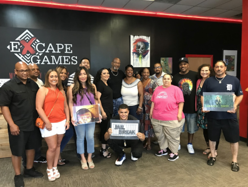 A diverse group of people poses together in an escape room venue, smiling and holding game signs.