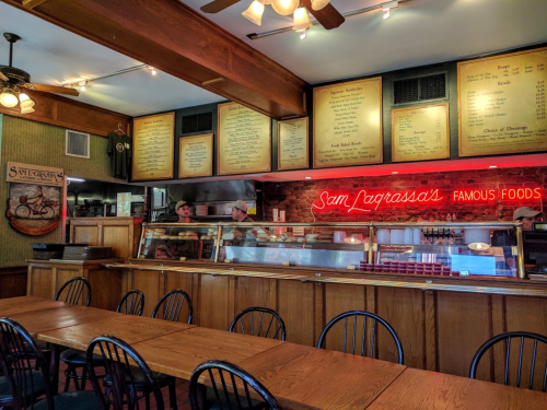 Interior of a cozy restaurant with wooden tables, a counter displaying food, and a neon sign reading "Sam Caprasso's Famous Foods."