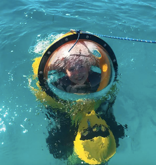 A child inside a clear underwater submersible, smiling while floating in turquoise water.