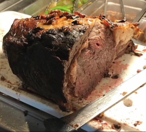 A large, roasted piece of meat with a crispy exterior, resting on a cutting board with juices around it.