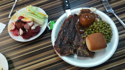 A plate with grilled steak, peas, mashed potatoes with gravy, a roll, and a side salad of mixed fruits and vegetables.