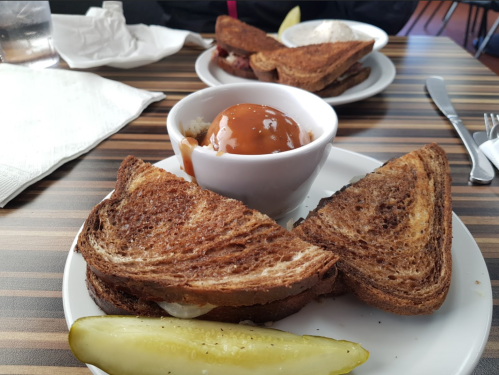 A plate with two toasted sandwiches, a bowl of caramel dessert, and a pickle on a wooden table.