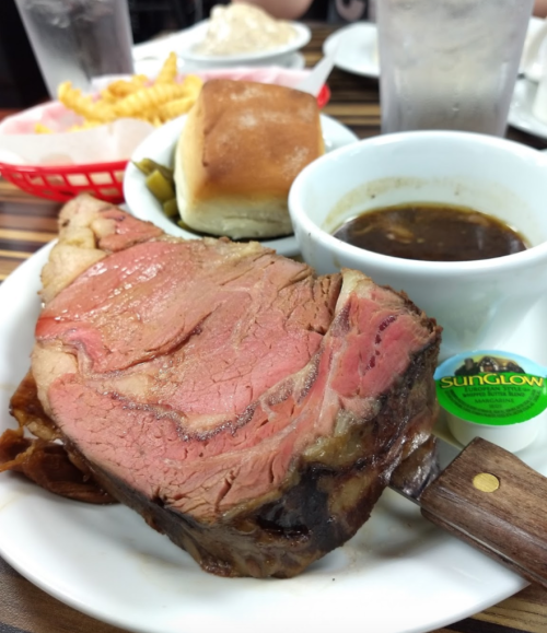 A plate featuring a large slice of prime rib, a roll, a cup of gravy, and pickles, with fries in the background.