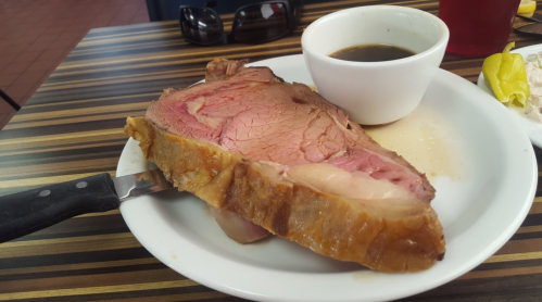 A plate with a large slice of prime rib, a small bowl of au jus, and a knife, set on a striped table.