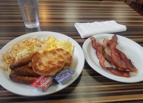 A plate of breakfast with scrambled eggs, hash browns, sausage links, a biscuit, and a side of crispy bacon.