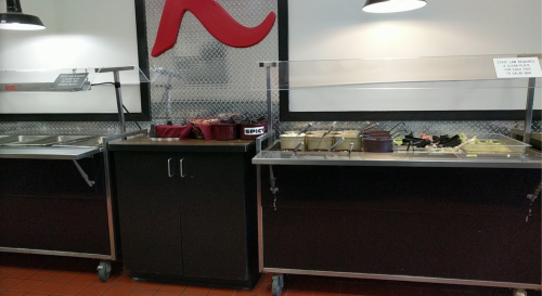 A food serving station with various trays and utensils, featuring a red logo on the wall behind.